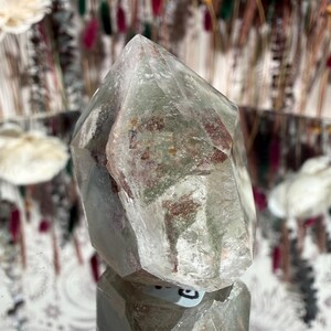 May include: A clear quartz crystal with green and reddish-brown inclusions. The crystal is faceted with a pointed top and sits on a reflective surface. The background features blurred floral arrangements.