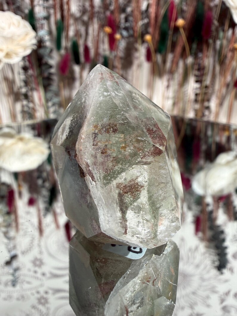 May include: A clear quartz crystal with green and reddish-brown inclusions. The crystal is faceted with a pointed top and sits on a reflective surface. The background features blurred floral arrangements.