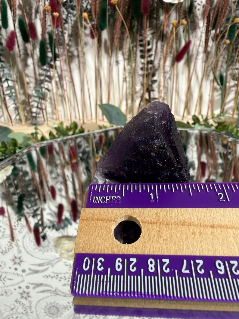 Puede incluir: Se muestra un cristal de amatista morado oscuro, de aproximadamente 3,8 cm de altura. Se coloca una regla de madera con marcas en pulgadas junto al cristal para comparar el tama&ntilde;o. El fondo presenta flores secas y una superficie reflectante.