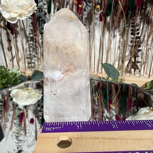 May include: A clear quartz crystal point with internal inclusions, displayed with a purple and wooden ruler. The crystal is approximately 3 inches tall. The background features dried flowers and a mirror.