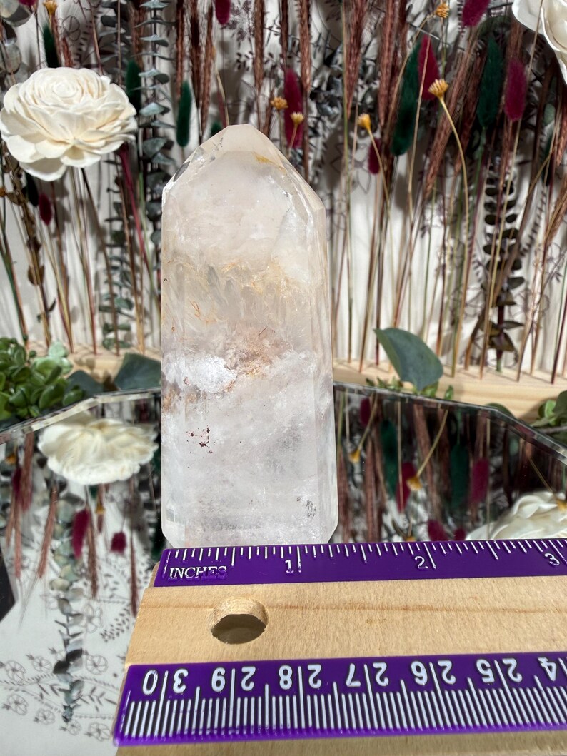 May include: A clear quartz crystal point with internal inclusions, displayed with a purple and wooden ruler. The crystal is approximately 3 inches tall. The background features dried flowers and a mirror.
