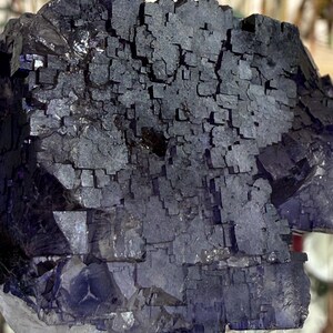 Puede incluir: Primer plano de un grupo de cristales de fluorita de color p&uacute;rpura oscuro. El cristal tiene una estructura c&uacute;bica con un color profundo, casi negro en algunas zonas. La base del cristal es de un p&uacute;rpura m&aacute;s claro y transl&uacute;cido.