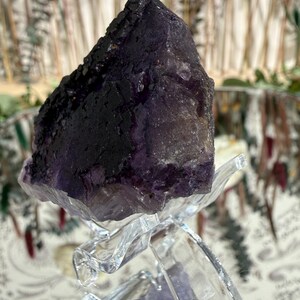 Puede incluir: Un cristal de fluorita morado oscuro, con una superficie rugosa y texturizada, se exhibe sobre un soporte decorativo transparente. El color del cristal var&iacute;a del violeta intenso a tonos m&aacute;s claros. El soporte es de pl&aacute;stico transparente moldeado.