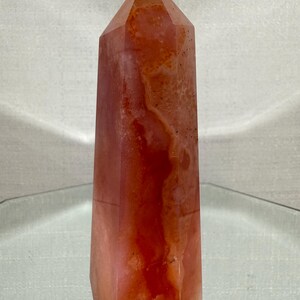 May include: A polished, six-sided crystal tower featuring a translucent, warm orange and reddish-pink hue. The stone displays natural internal cloudiness, subtle mineral inclusions, and a smooth, faceted surface that tapers to a point.