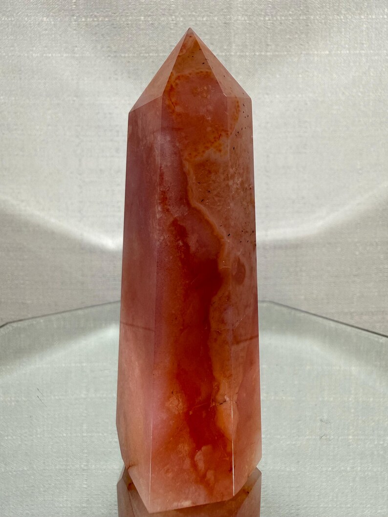 May include: A polished, six-sided crystal tower featuring a translucent, warm orange and reddish-pink hue. The stone displays natural internal cloudiness, subtle mineral inclusions, and a smooth, faceted surface that tapers to a point.