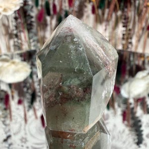 May include: A faceted quartz crystal with green and reddish inclusions. The crystal is transparent with a pointed top and sits on a reflective surface. The background features blurred floral arrangements.