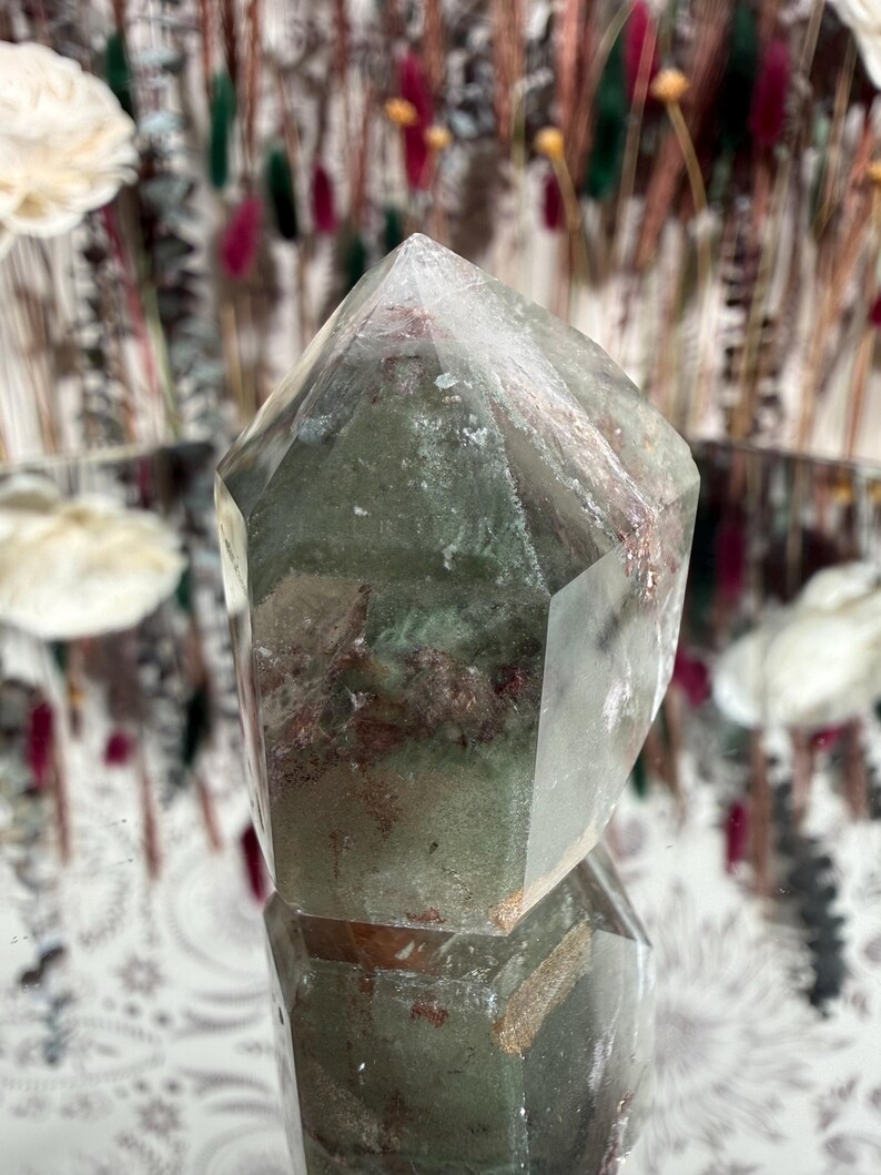 May include: A faceted quartz crystal with green and reddish inclusions. The crystal is transparent with a pointed top and sits on a reflective surface. The background features blurred floral arrangements.