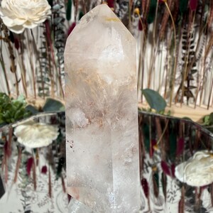 May include: A clear quartz crystal point with internal inclusions of brown and orange hues. The crystal is faceted and tapers to a point. The background features dried flowers and a decorative mirror.