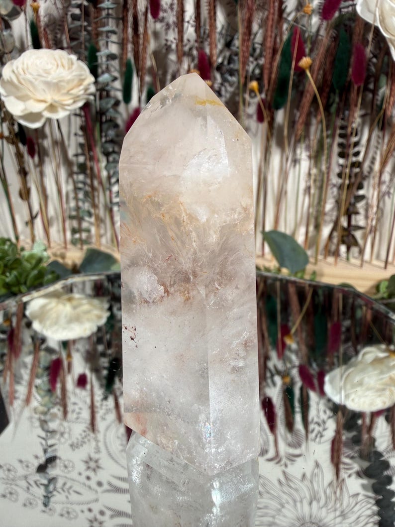 May include: A clear quartz crystal point with internal inclusions of brown and orange hues. The crystal is faceted and tapers to a point. The background features dried flowers and a decorative mirror.