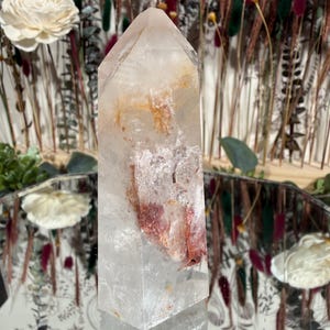 May include: A clear quartz crystal tower with reddish-brown and yellow inclusions. The crystal is faceted and stands upright, reflecting light. The background includes dried flowers and decorative elements, suggesting a focus on natural decor and spiritual items.