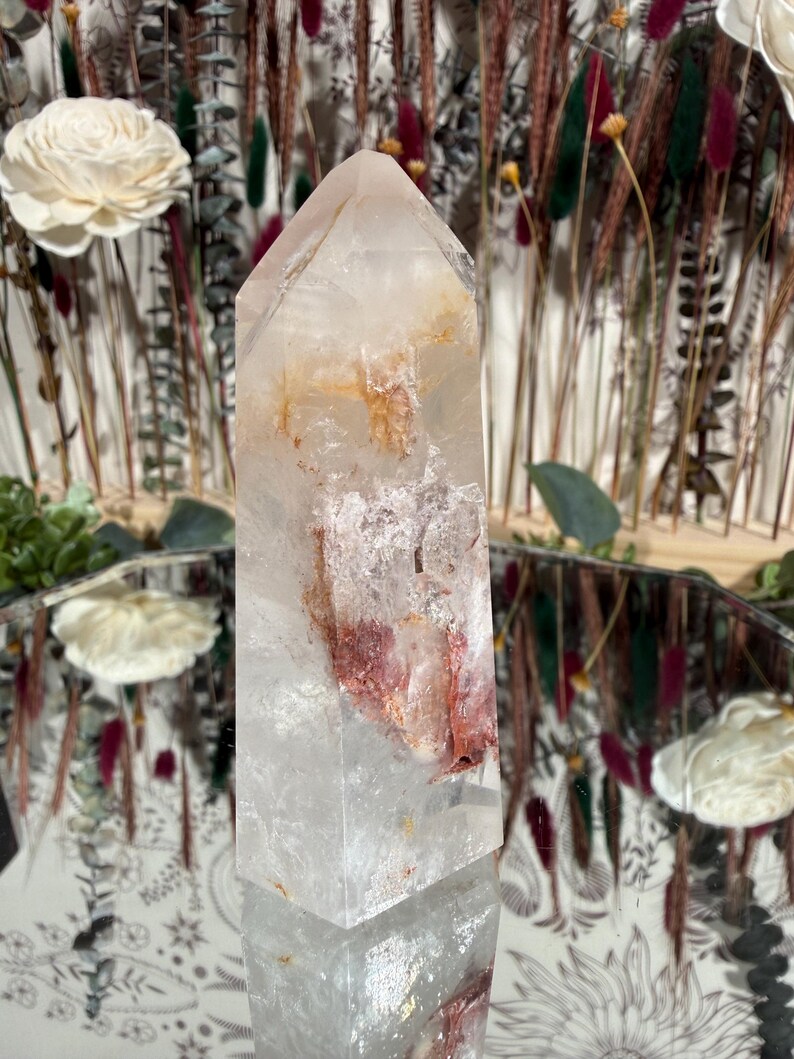 May include: A clear quartz crystal tower with reddish-brown and yellow inclusions. The crystal is faceted and stands upright, reflecting light. The background includes dried flowers and decorative elements, suggesting a focus on natural decor and spiritual items.