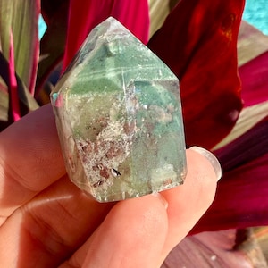May include: A faceted crystal tower with green and brown inclusions. The crystal is held in a hand, with a background of red and green leaves and a glimpse of blue water.