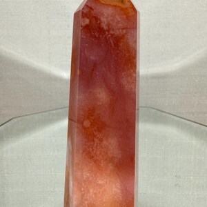May include: A polished, six-sided crystal tower featuring a translucent, warm orange and reddish-pink colour palette. The stone displays natural internal cloudiness, subtle white inclusions, and deep amber-coloured veins throughout its vertical structure.