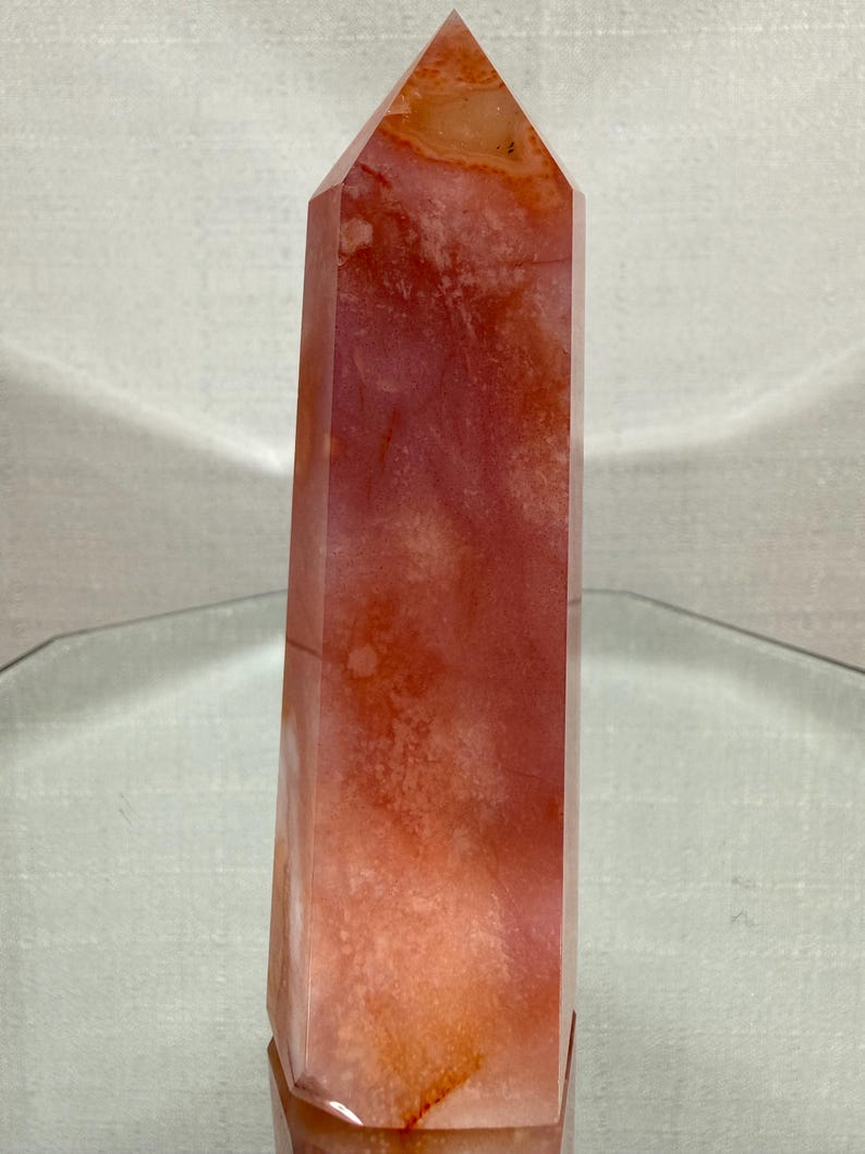 May include: A polished, six-sided crystal tower featuring a translucent, warm orange and reddish-pink colour palette. The stone displays natural internal cloudiness, subtle white inclusions, and deep amber-coloured veins throughout its vertical structure.