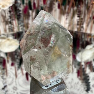 May include: A clear, faceted crystal point with internal inclusions of green and red. The crystal is displayed on a reflective surface, showcasing its geometric shape and natural variations. The background features decorative elements.