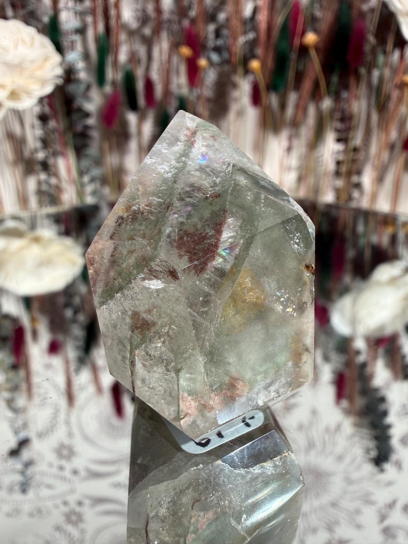 May include: A clear, faceted crystal point with internal inclusions of green and red. The crystal is displayed on a reflective surface, showcasing its geometric shape and natural variations. The background features decorative elements.
