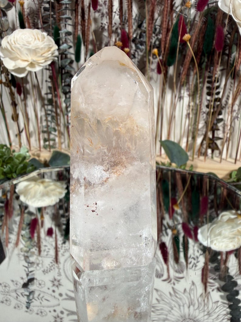 May include: A clear quartz crystal point with internal inclusions of tan and brown hues. The crystal is faceted and stands upright, reflecting light. The background features dried flowers and decorative elements, suggesting a focus on natural decor and spiritual items.