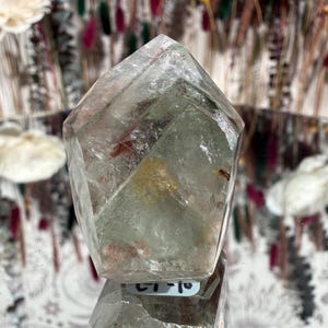 May include: A clear quartz crystal with a faceted, geometric shape. The crystal has internal inclusions of green and red hues. The crystal is displayed on a reflective surface, with a blurred background of dried flowers.