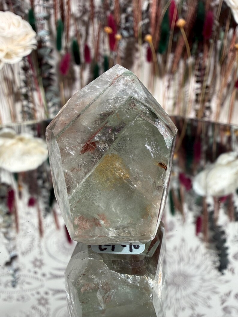 May include: A clear quartz crystal with a faceted, geometric shape. The crystal has internal inclusions of green and red hues. The crystal is displayed on a reflective surface, with a blurred background of dried flowers.
