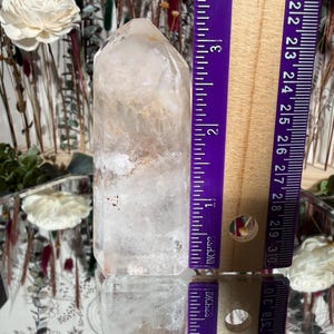 May include: A clear quartz crystal point, approximately 3 inches (7.6 cm) tall, with internal inclusions and a slightly rounded tip. The crystal is positioned vertically, reflecting light and set against a blurred background with floral elements.