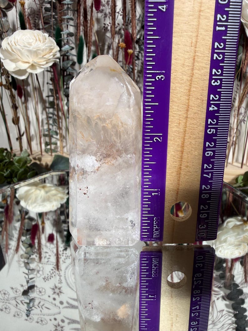 May include: A clear quartz crystal point, approximately 3 inches (7.6 cm) tall, with internal inclusions and a slightly rounded tip. The crystal is positioned vertically, reflecting light and set against a blurred background with floral elements.