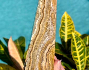 May include: A polished crystal tower featuring horizontal bands of golden yellow, brown, and white mineral layers. The stone has a pointed top and a vertical fissure running through the center, revealing a textured, crystalline interior structure.