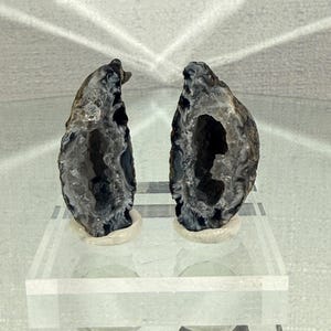 May include: Two halves of a split agate geode standing upright on small circular bases. The exterior of the stones is dark and textured, while the interior reveals a hollow cavity lined with translucent, crystalline formations in shades of gray.
