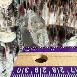 May include: A clear quartz crystal with green and red inclusions. The crystal is faceted and stands upright. A purple ruler with inch measurements is in the foreground, showing the crystal's size. The background features dried flowers and a mirror.