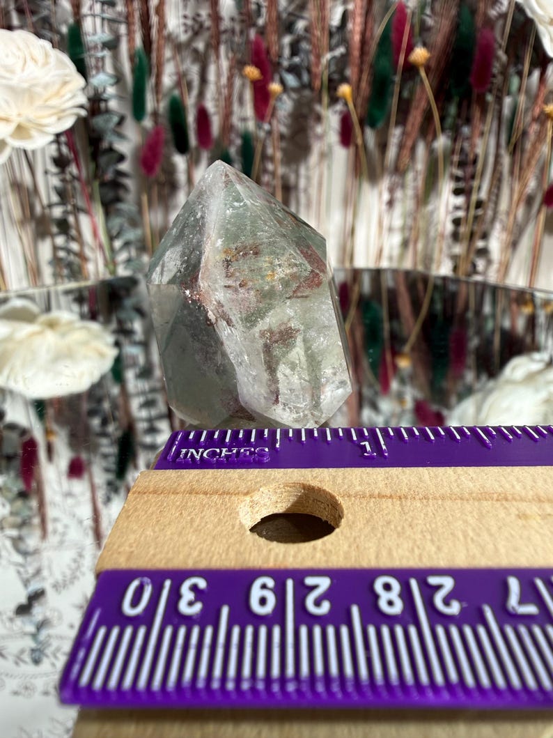May include: A clear quartz crystal with green and red inclusions. The crystal is faceted and stands upright. A purple ruler with inch measurements is in the foreground, showing the crystal's size. The background features dried flowers and a mirror.
