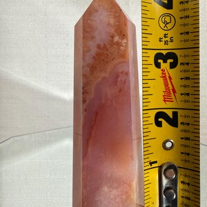 May include: A polished, multi-faceted crystal tower featuring translucent shades of peach, coral, and cream with internal dendritic inclusions. A yellow Milwaukee measuring tape placed alongside indicates the crystal stands 10.16 cm tall.