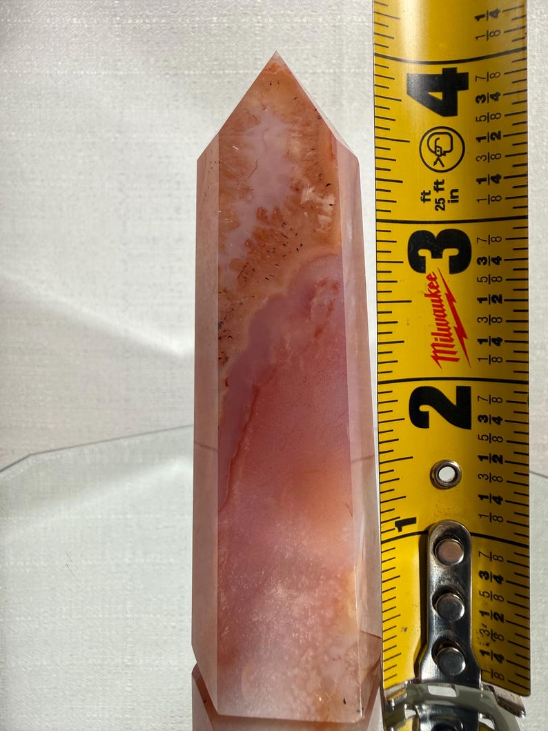 May include: A polished, multi-faceted crystal tower featuring translucent shades of peach, coral, and cream with internal dendritic inclusions. A yellow Milwaukee measuring tape placed alongside indicates the crystal stands 10.16 cm tall.