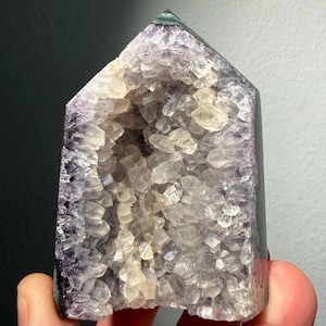 May include: A polished amethyst crystal point with a hollow center filled with translucent, jagged quartz crystals. The exterior of the stone is a deep purple, while the interior cavity features clear, light-colored crystalline structures.