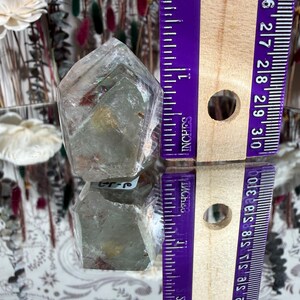 May include: A clear quartz crystal point, approximately 1.5 inches tall, with internal inclusions and a reflective surface. The crystal is displayed next to a purple ruler marked in inches. The crystal is on a reflective surface.