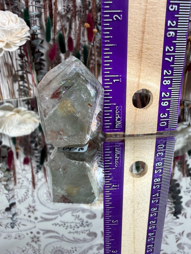 May include: A clear quartz crystal point, approximately 1.5 inches tall, with internal inclusions and a reflective surface. The crystal is displayed next to a purple ruler marked in inches. The crystal is on a reflective surface.