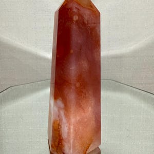 May include: A polished crystal tower with a hexagonal base and a pointed top. The stone features a translucent appearance with swirling patterns of deep orange, burnt red, and creamy white hues throughout its smooth, multifaceted vertical surface.