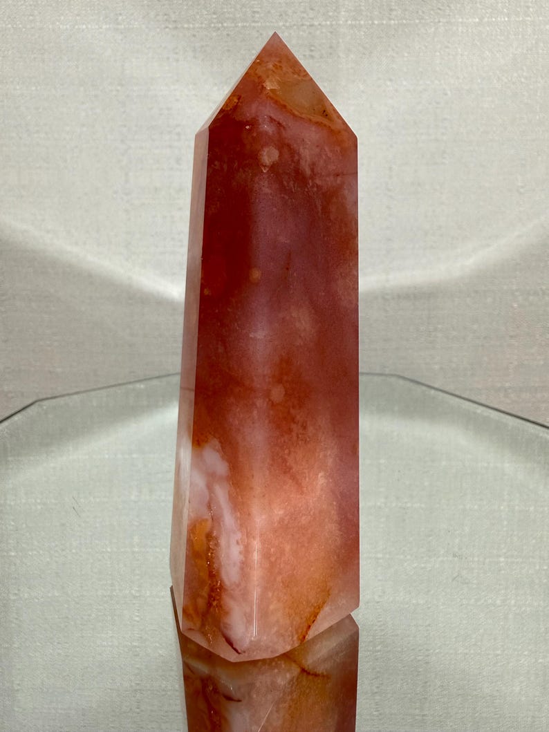 May include: A polished crystal tower with a hexagonal base and a pointed top. The stone features a translucent appearance with swirling patterns of deep orange, burnt red, and creamy white hues throughout its smooth, multifaceted vertical surface.