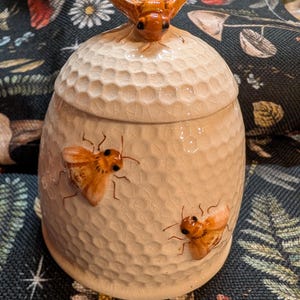Puede incluir: Un tarro de miel de cerámica con tapa, con una superficie texturizada y adornos de abejas tridimensionales. El tarro es de color blanquecino con abejas marrones, y la tapa está coronada con una abeja más grande. El tarro tiene un patrón de panal.