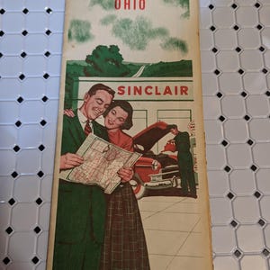 Può includere: Un opuscolo vintage con mappa stradale Sinclair Ohio che presenta un'illustrazione a colori di una coppia che guarda una mappa davanti a una stazione di servizio con un addetto che lavora su un'auto rossa. Il testo OHIO e SINCLAIR è stampato.