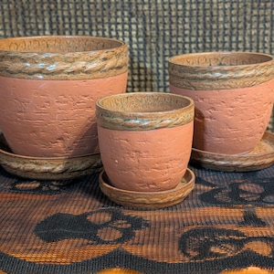 Może przedstawiać: Trzy ceramiczne donice w kolorze terakoty o różnych rozmiarach, każda z teksturowaną powierzchnią i ozdobnym, sznurowatym pasem wokół krawędzi. Każda donica stoi na pasującym spodku. Nadaje się do użytku wewnątrz i na zewnątrz.