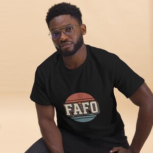 Puede incluir: Camiseta negra con un gráfico retro. El diseño presenta la palabra "FAFO" en una fuente vintage audaz, dentro de un diseño circular con rayas naranjas y azules. La camiseta está hecha de un material suave.