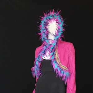 May include: A mannequin wearing a vibrant pink jacket with gold detailing and blue and purple faux fur trim. The jacket is styled with a black dress and a matching fur hood and scarf, set against a black backdrop.