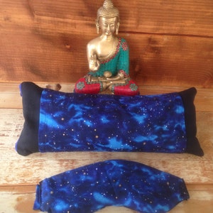 May include: A blue and black eye pillow and eye mask set with a celestial pattern of white and gold stars. The pillow is rectangular, and the mask is curved. A golden Buddha statue sits in the background.