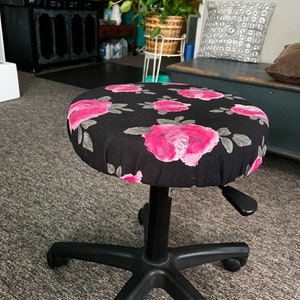 May include: A black office chair with a round seat covered in a fabric with a pink rose pattern. The chair has a black base with five wheels and a lever for adjusting the height.