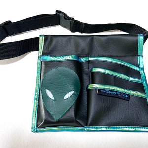 May include: Black fanny pack with a green alien face applique. The pack has multiple pockets and a black adjustable strap. The edges are trimmed with iridescent teal material. The text "The Escape Portal" is on a small black tag.