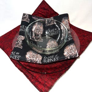 May include: A clear glass bowl sits atop a clear glass plate, with a black and white patterned fabric featuring the text "GAME OF THRONES" and a throne design. A red patterned fabric square is underneath.