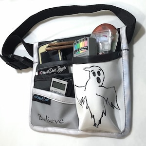 May include: A white and grey utility bag with a black strap, featuring multiple pockets. The bag contains various ghost hunting tools, including a meter, a device with a colour scale, and a small digital device. A ghost is drawn on the front, and the word "Believe" is written on the bag.