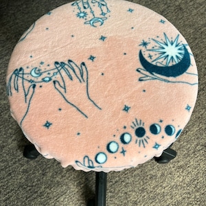 May include: A round, pink stool cover with a celestial design. The cover features teal illustrations of hands, moons, stars, and phases of the moon. The stool has a black base and is set on a neutral-colored carpet.