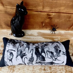 May include: A rectangular pillow with black end panels and a gray center panel featuring a black cat print. A glossy black cat figurine and a black plastic spider are also in the image. The pillow is a Halloween-themed item.