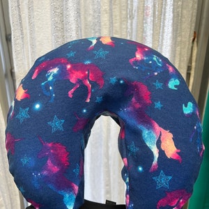 May include: A navy blue travel pillow with a U-shape design. The pillow features a pattern of colorful unicorns, stars, and a galaxy design. The pillow is made of soft fabric and is designed for neck support.