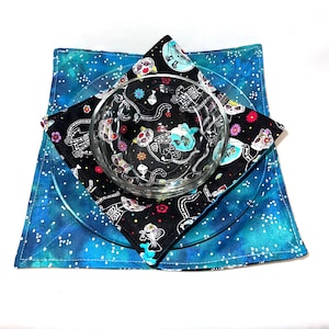 May include: A clear glass bowl and plate set atop a black and blue patterned placemat. The placemat features a Day of the Dead cat design. The plate and placemat are on a blue and white speckled background.